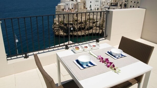 Un balcón con una mesa lista para dos personas, con vistas a un pueblo costero, edificios y el mar.