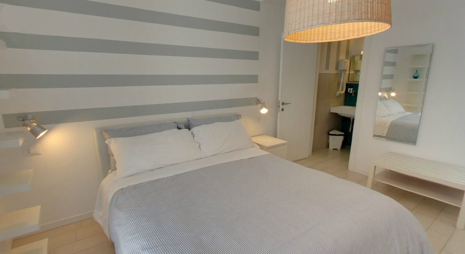A bedroom with a bed, bedside lamps, a hanging light fixture, a mirror, and a bathroom visible through an open door.