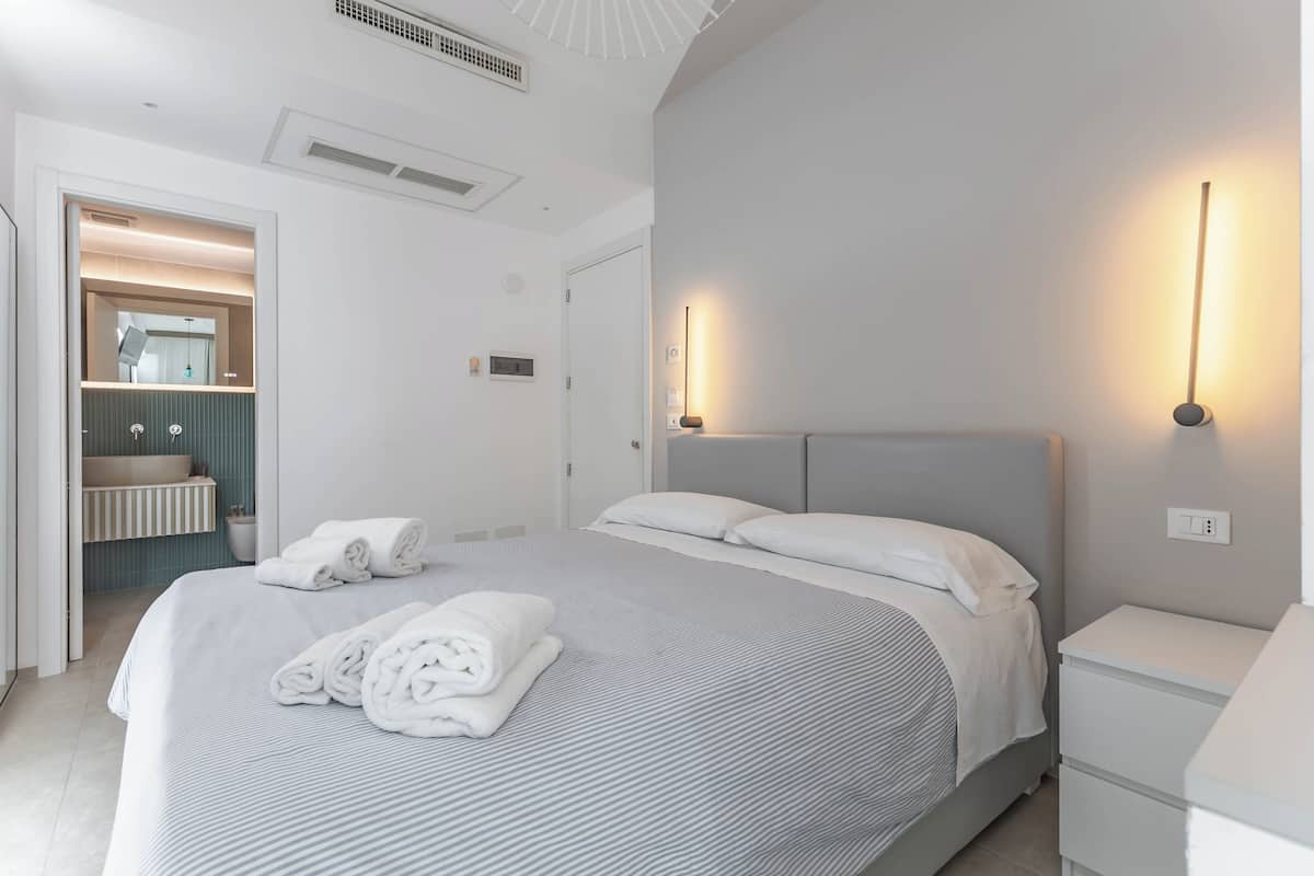 A modern hotel room with a large bed, a bedside table, a nightstand, a built-in vanity with a mirror, and a towel rack.