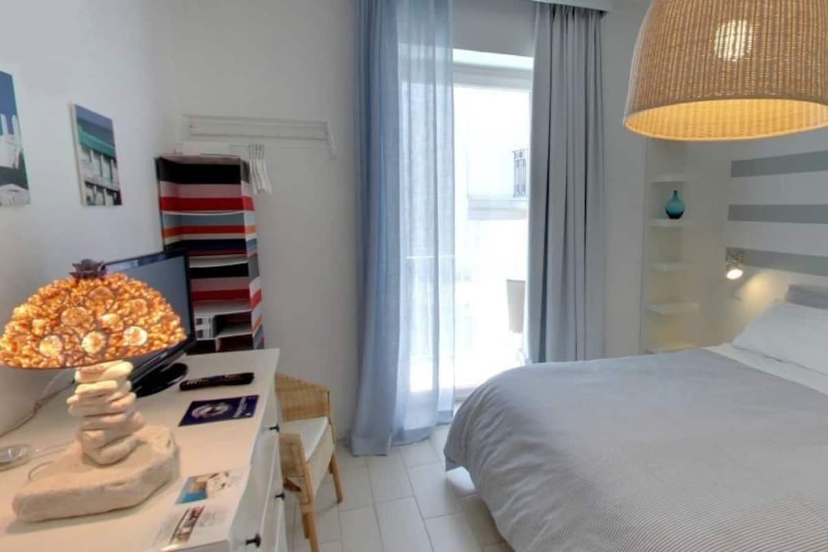 A bedroom with a bed, a bedside table with a lamp, a window with curtains, and a striped wall.