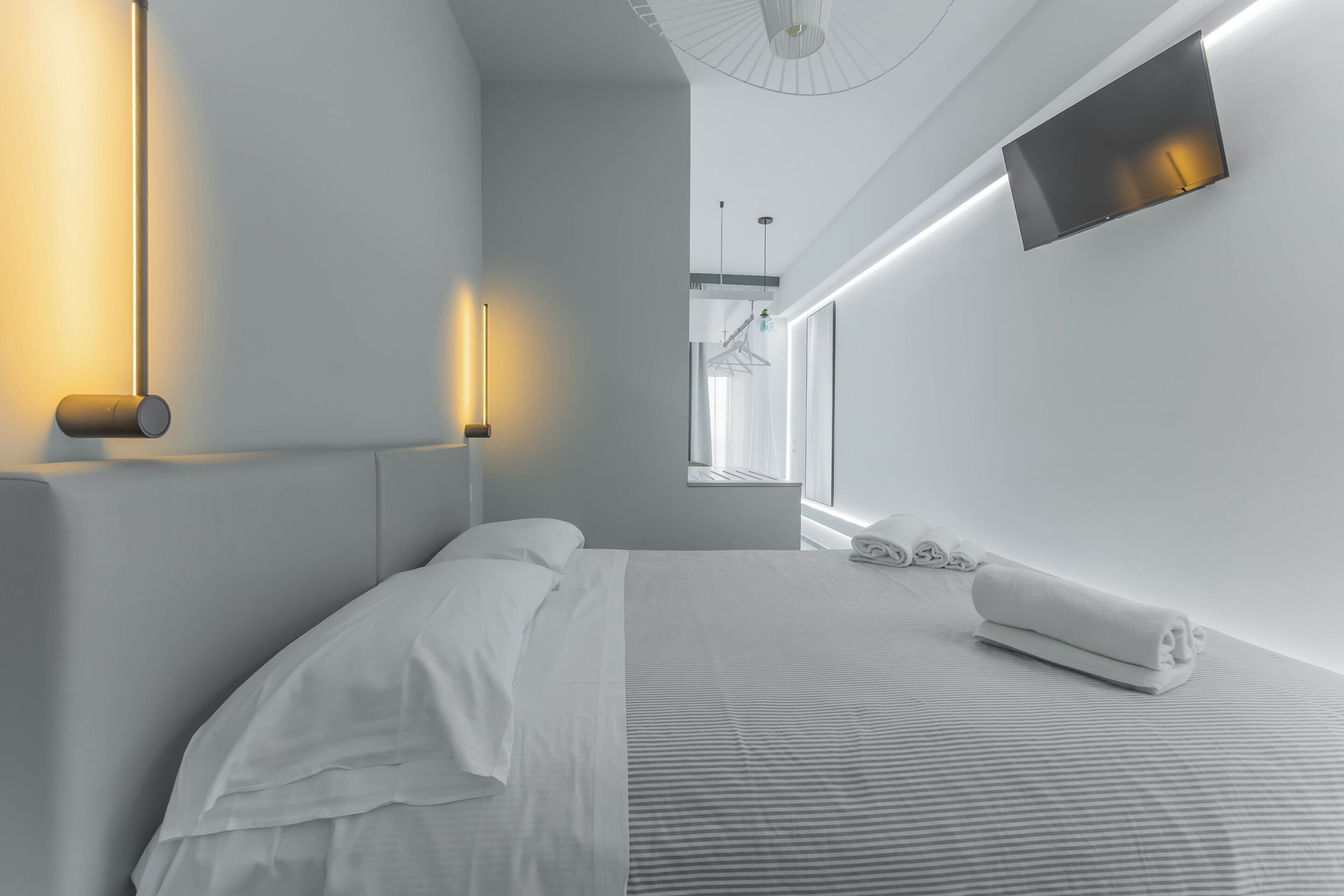A modern hotel room with a neatly made bed, a flat-screen TV mounted on the wall, and a large window providing natural light.