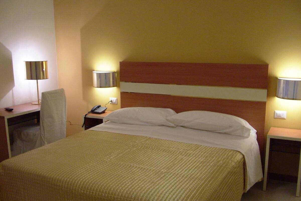Ein Hotelzimmer mit einem großen Bett, einem Schreibtisch mit Lampe, einem Sessel und einem Telefon auf dem Nachttisch.