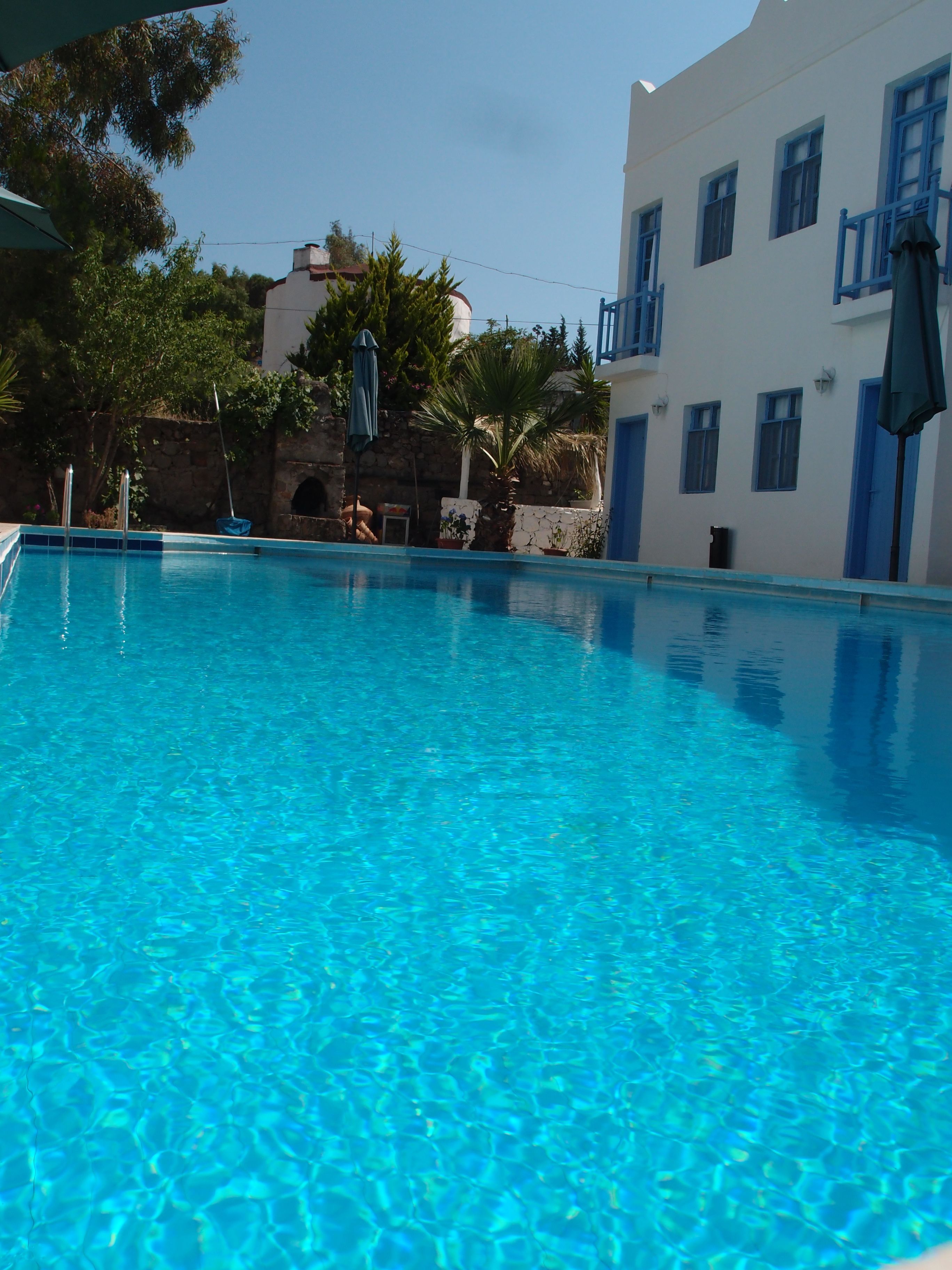 A clear blue swimming pool surrounded by white buildings with blue accents, a stone wall, and some greenery.