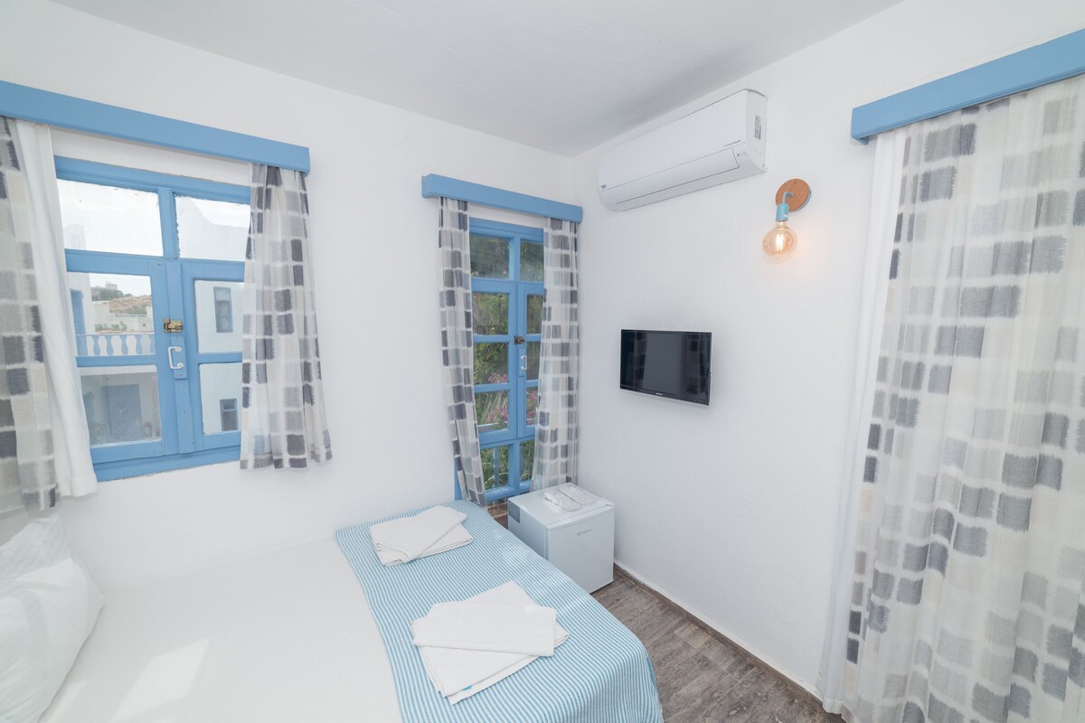 Un dormitorio con cama, mesita de noche, televisor y ventana con cortinas.