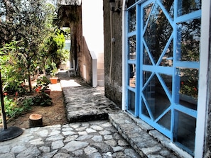 Stroll through the serene garden, surrounded by lush greenery and potted plants, to a charming stone building with a distinctive blue window.