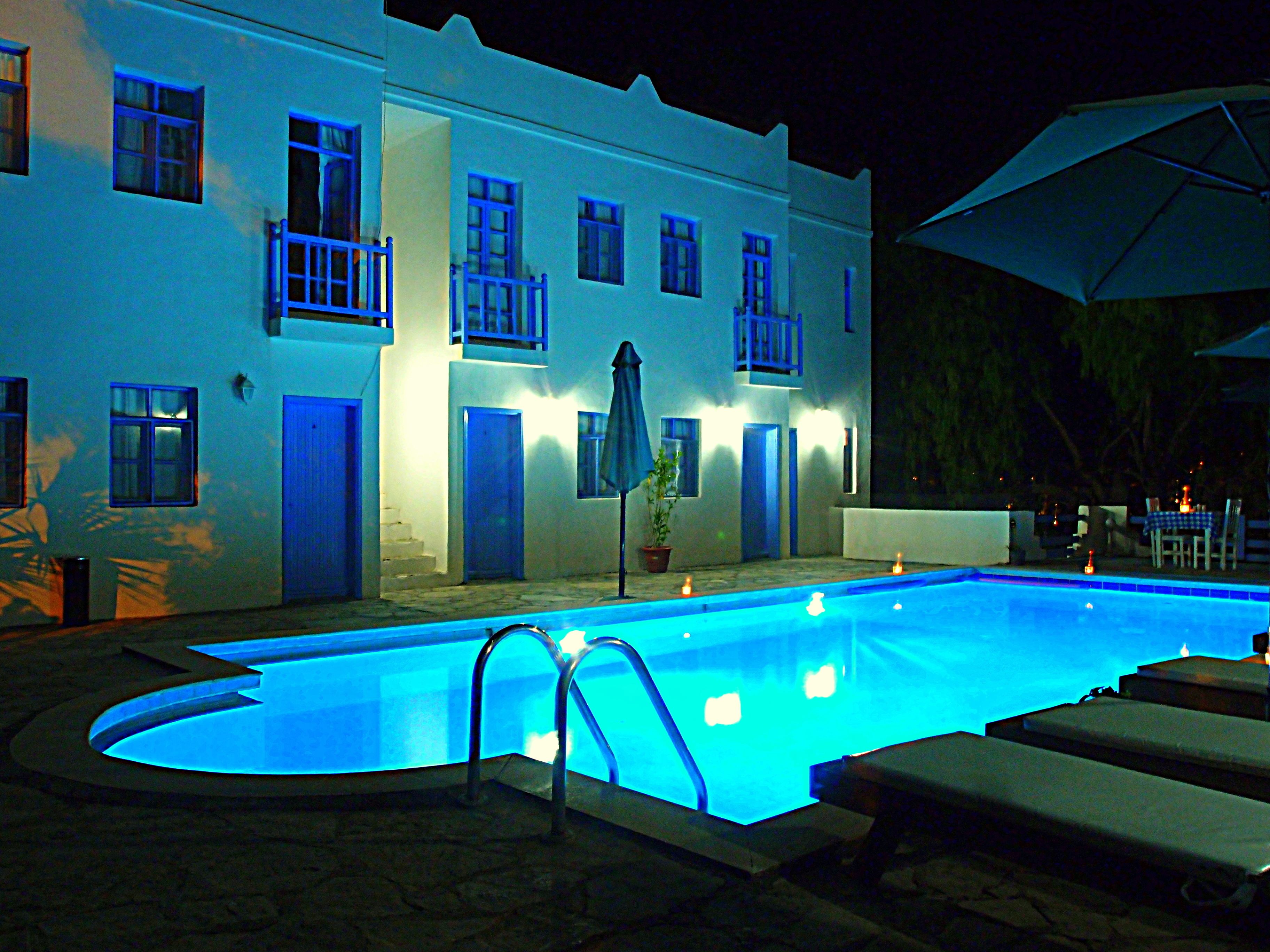 A nighttime swimming pool area with a white building, blue balconies, and a covered seating area.