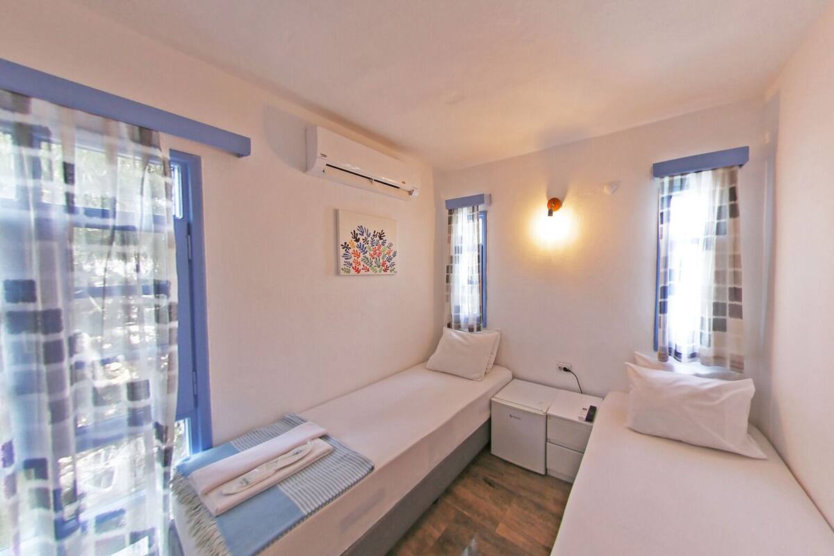 A small, well-lit room with two beds, a desk, and an air conditioning unit.