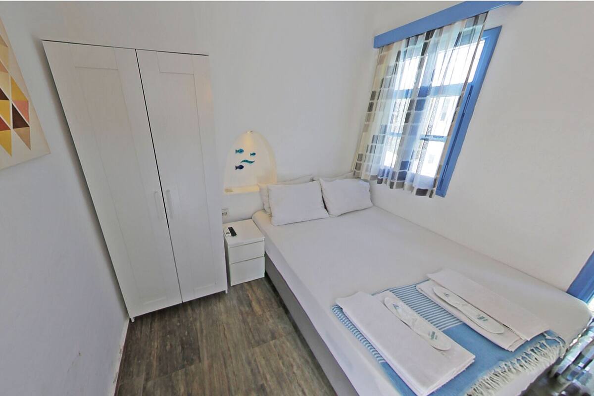 A small, single-bed room with a wooden floor, a white wardrobe, a bedside table with a lamp, and a window with curtains.