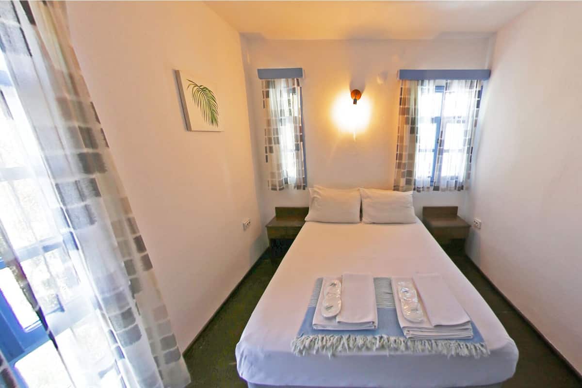 A small, single-bed room with a bed at the center, two windows with curtains, a wall-mounted light fixture, and a small painting on the wall.