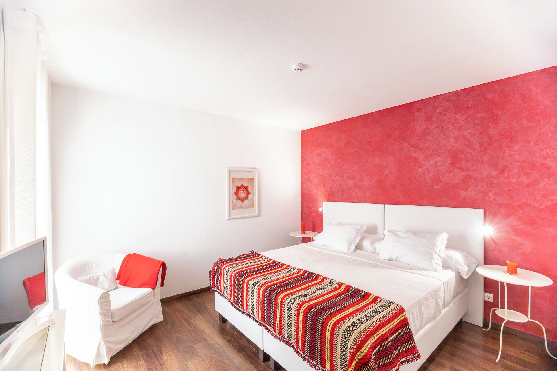 A modern bedroom with a large bed, a red accent wall, a white headboard, and a small bedside table with a lamp.