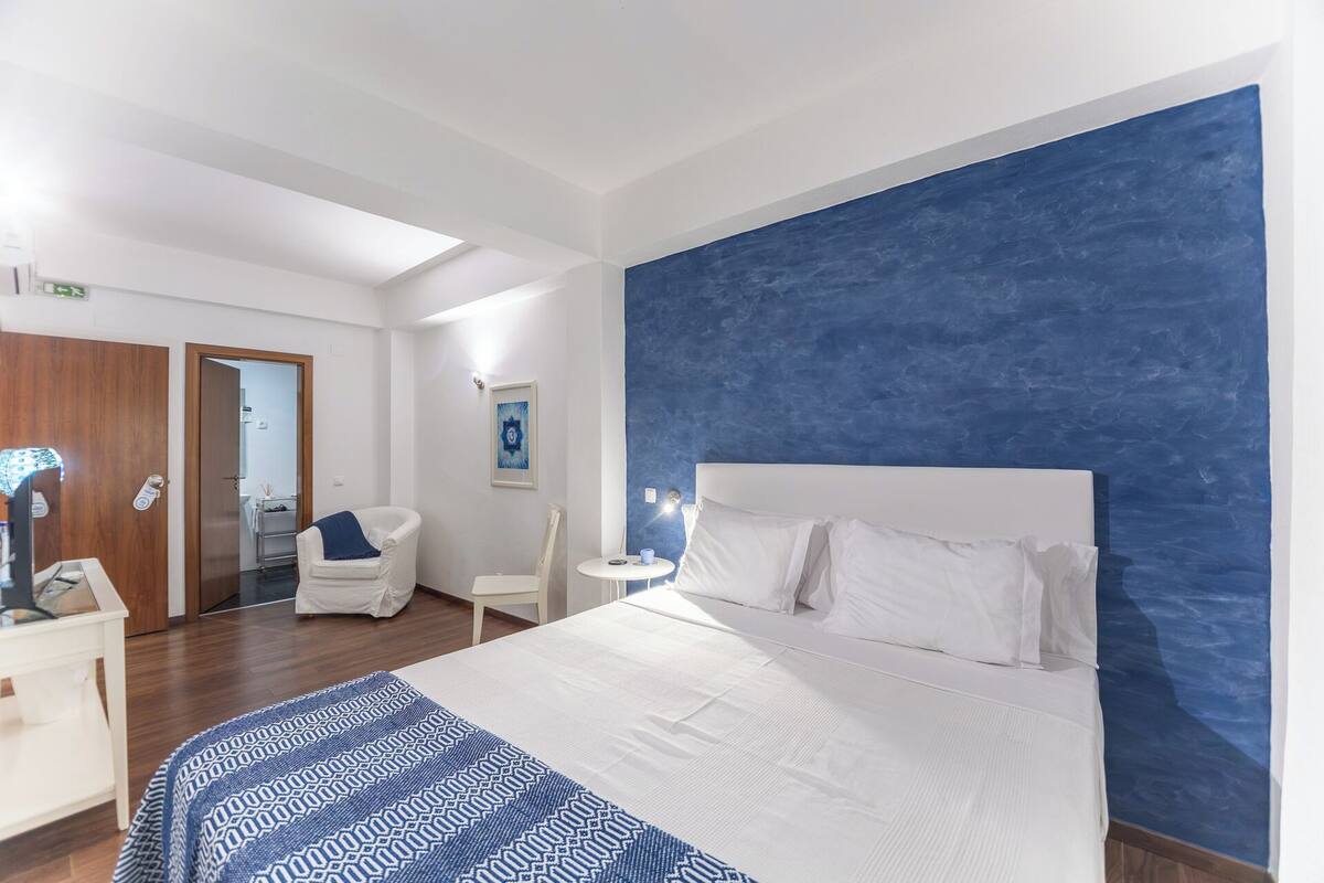 A hotel room with a large bed, a white chair, a small round table, and a blue accent wall.