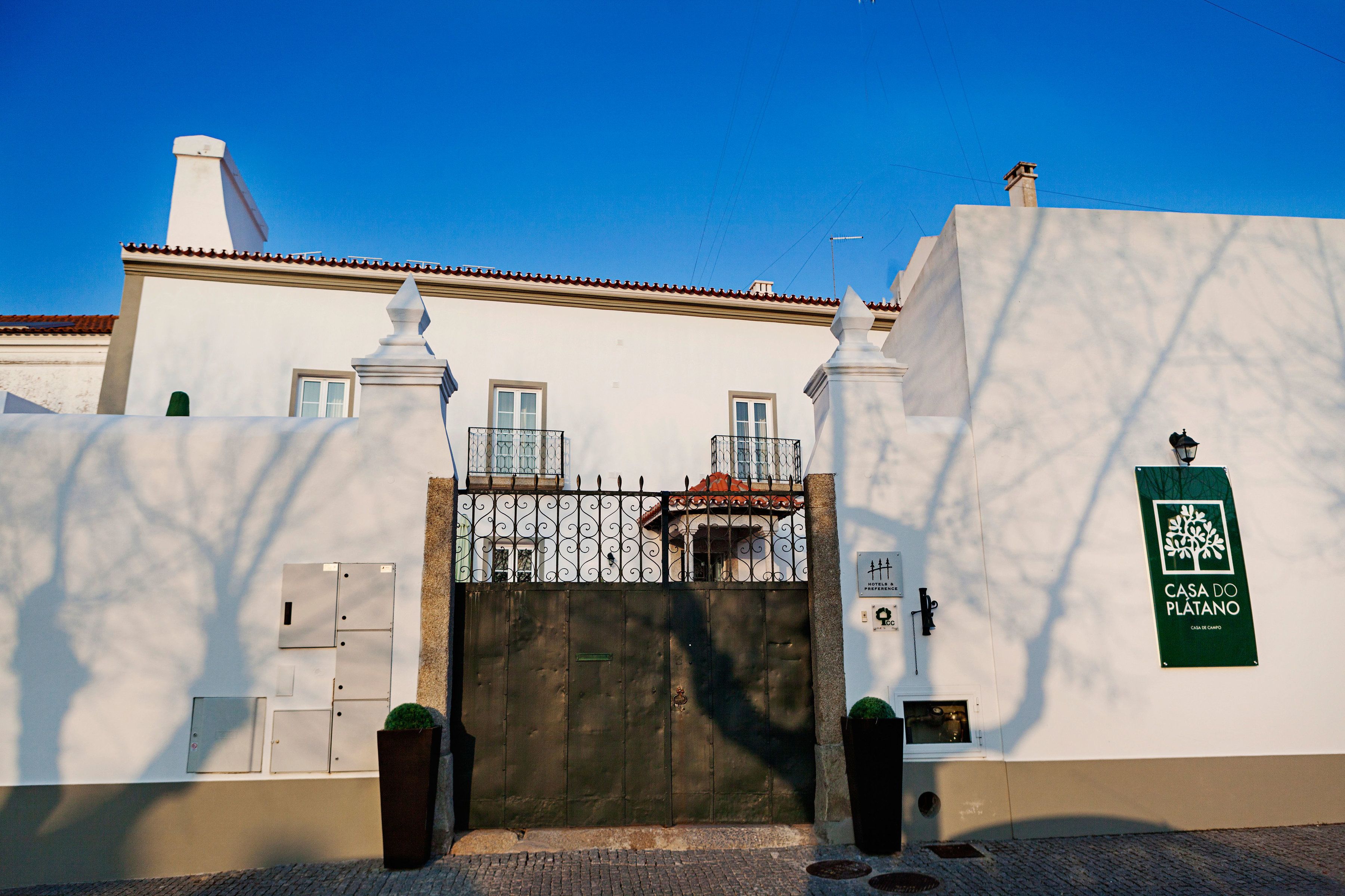 Uma entrada protegida por um portão de metal, um edifício branco com telhado em telhas e uma placa verde na parede.