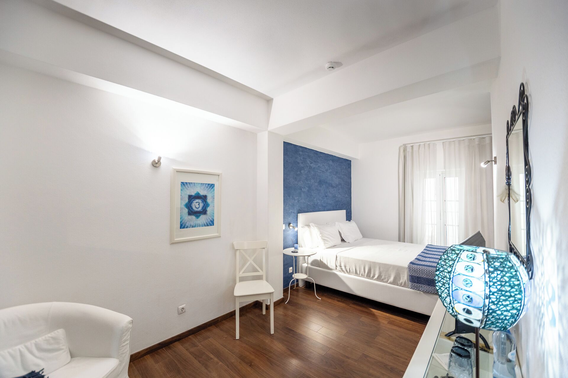 A hotel room with a bed, a chair, a small table, and a painting on the wall.