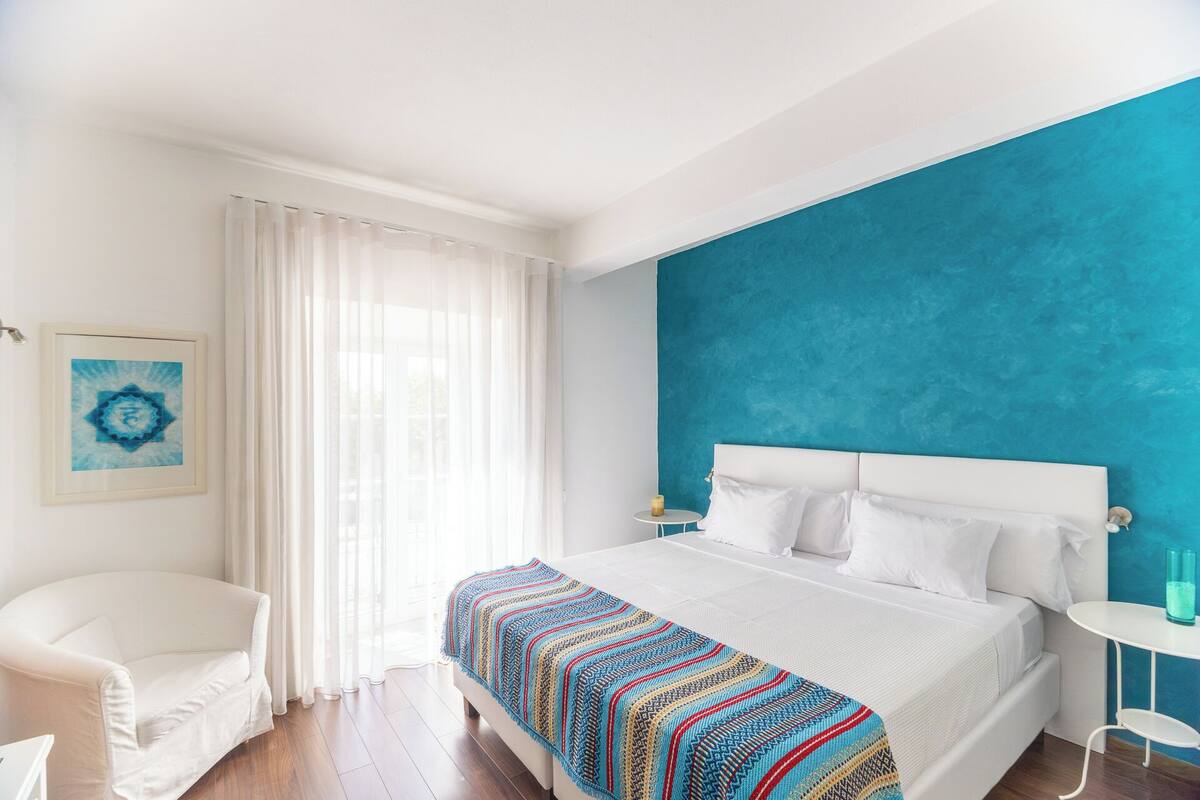 A bedroom with a large bed, white curtains, a blue accent wall, and a framed artwork on the wall.