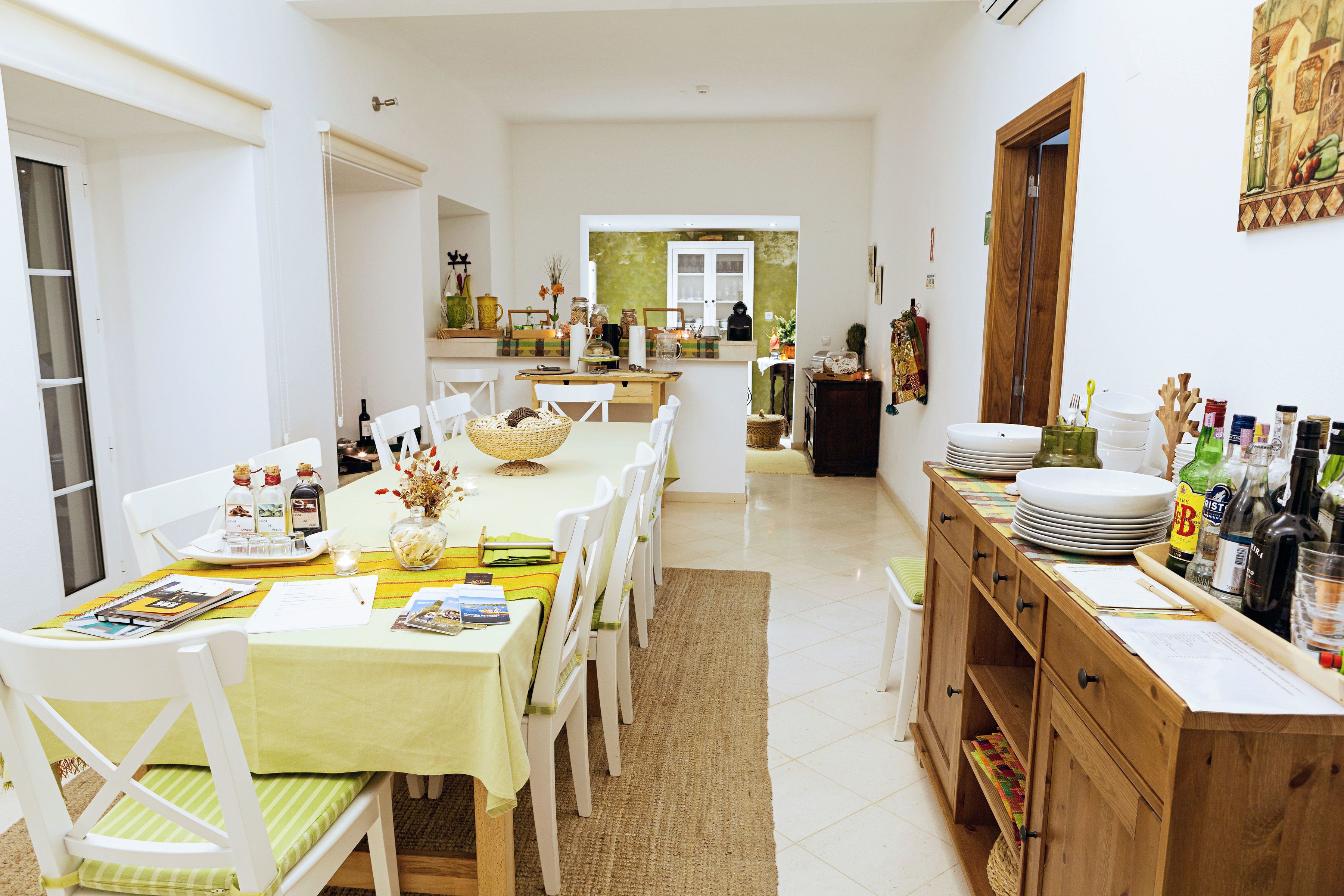 A spacious kitchen with a dining area, featuring a long table, chairs, and a wooden cabinet with kitchenware.