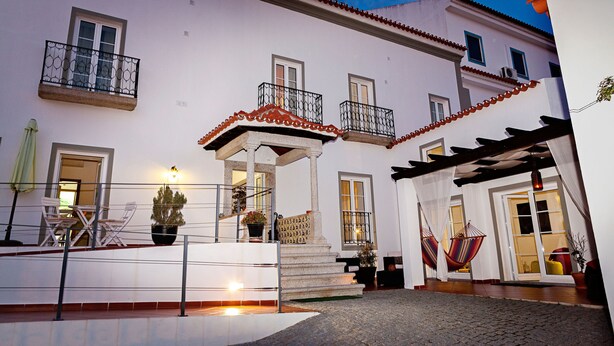 A white building with balconies, a covered area with chairs, and a cobblestone path. - Casa Do Platano (Arraiolos)