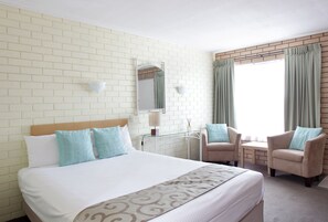 Habitación de hotel con una cama grande, dos sillones, una mesita, un espejo y una ventana con cortinas.