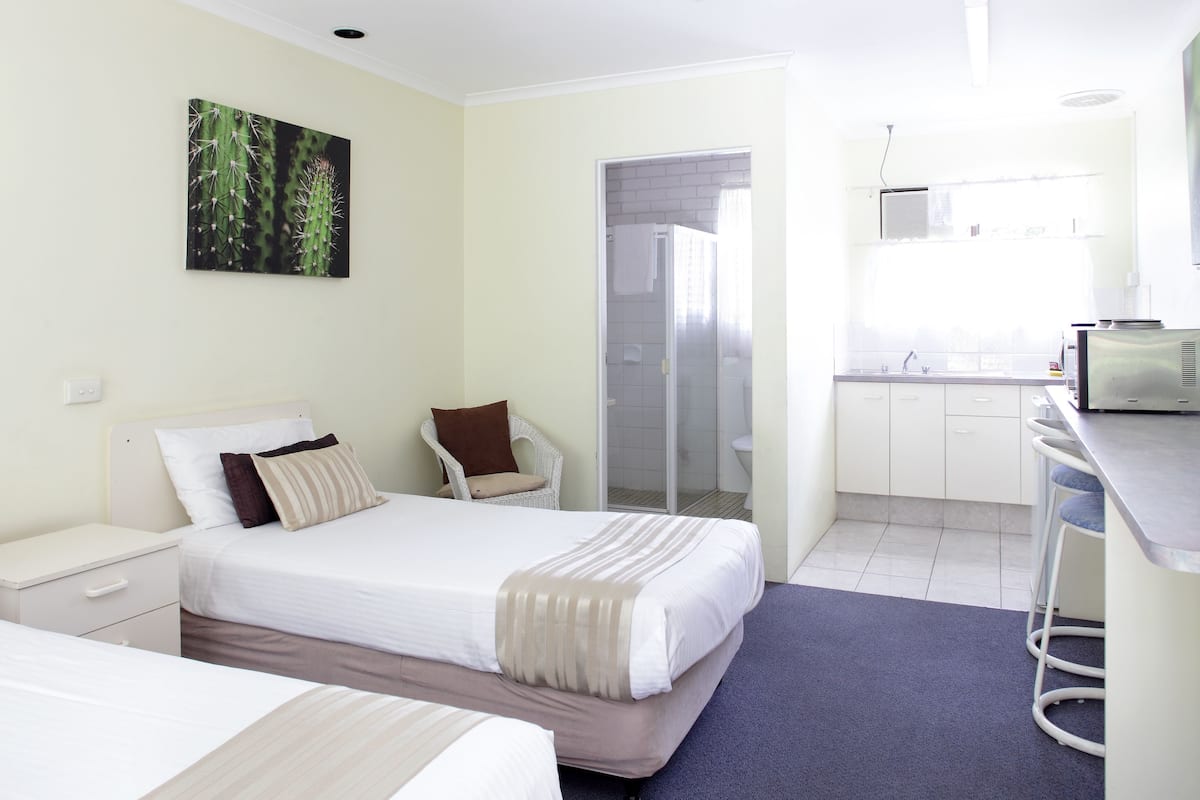 A hotel room with two beds, a small seating area, a kitchenette, and a shower area.