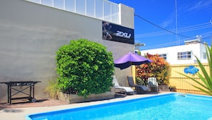 Una piscina con borde azul, un área de patio con mesa y sillas, y un arbusto grande.