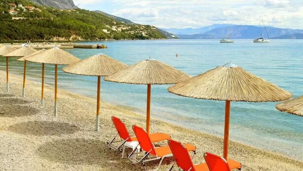 A beach with clear water, a row of orange beach chairs, and straw umbrellas. - Comfy Hostel Studios (Corfu)