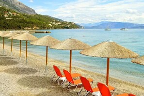 A beach with clear water, a row of orange beach chairs, and straw umbrellas. - Comfy Hostel Studios (Corfu)