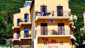 A two-story hotel with balconies, a sign indicating it is a hostel, and a mountainous backdrop. - Comfy Hostel Studios (Corfu)