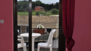 A balcony with a table and chairs, a red curtain, and a view of a building and greenery. - Comfy Hostel Studios (Corfu)