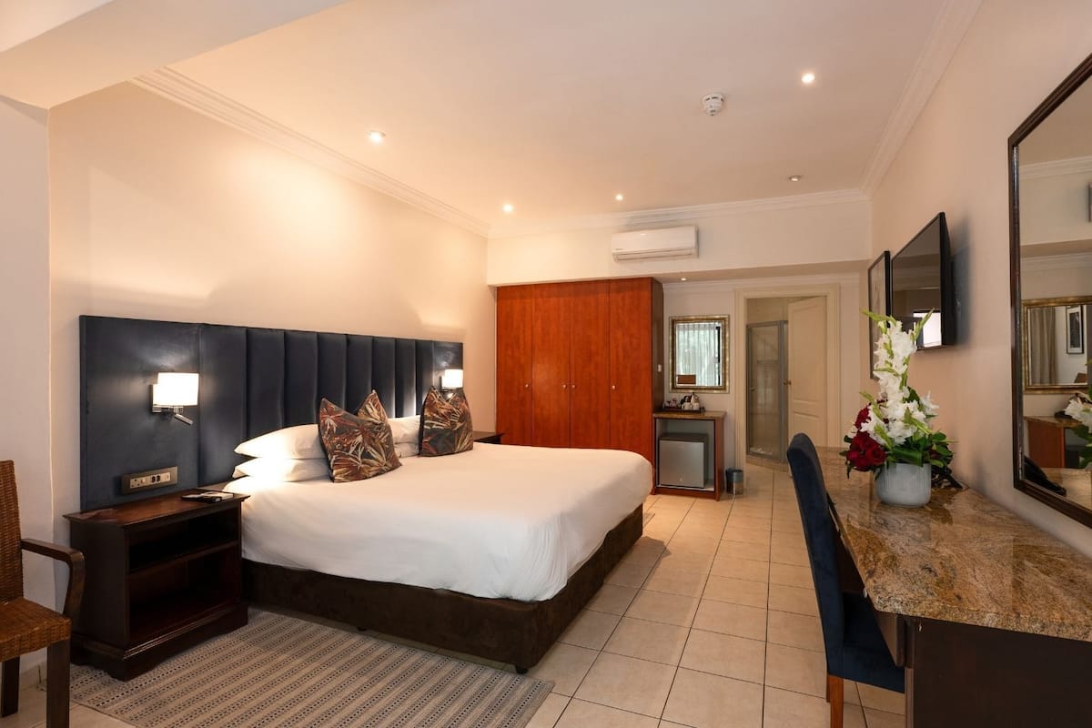 A hotel room with a large bed, bedside tables, a desk, a chair, a mirror, and an air conditioning unit.