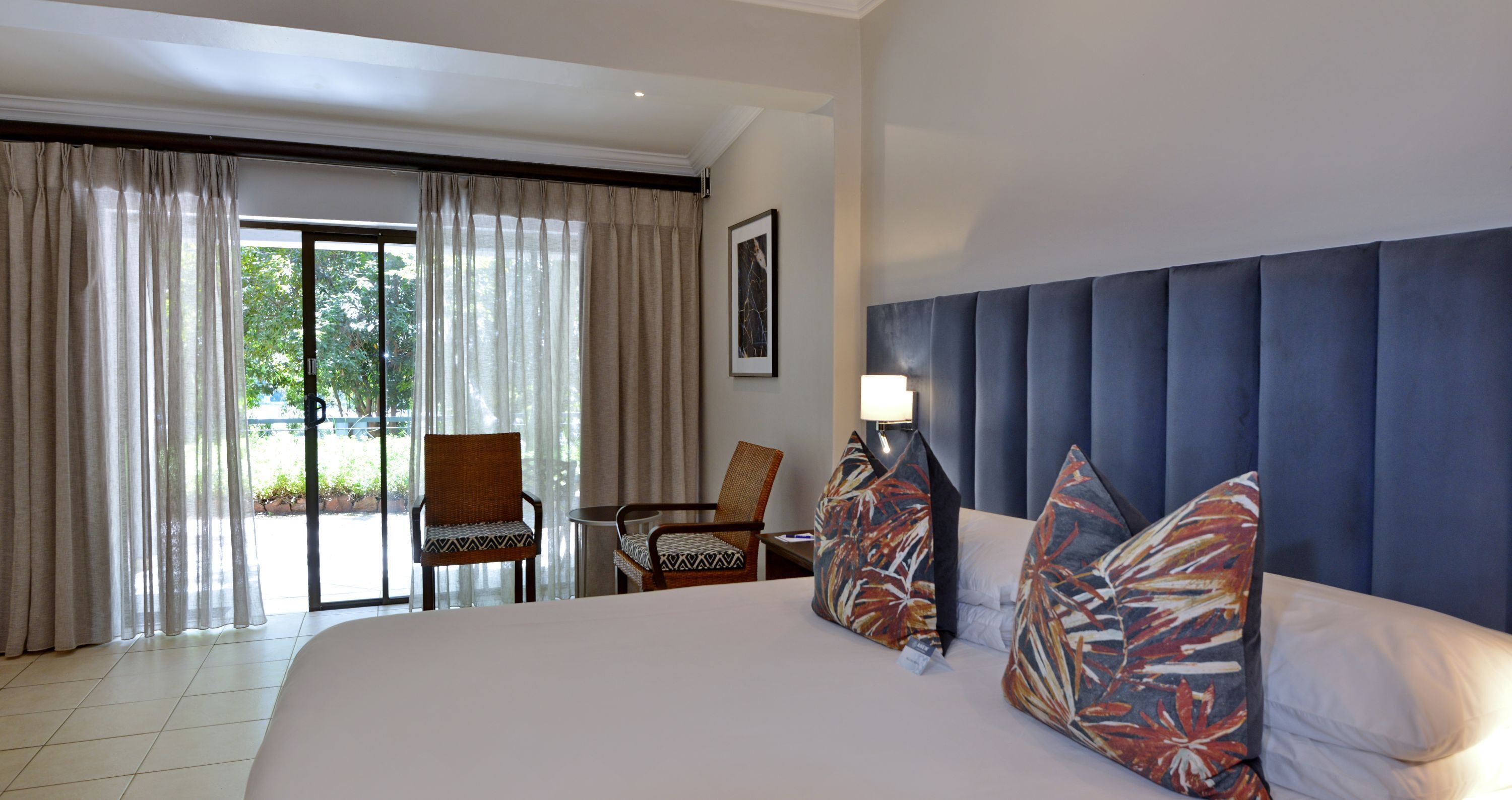 A hotel room with a large bed, two chairs, a table, and a view of the outdoors through a sliding door.