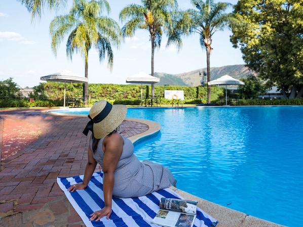 A person sitting by a pool with a towel and a book, surrounded by palm trees and umbrellas.