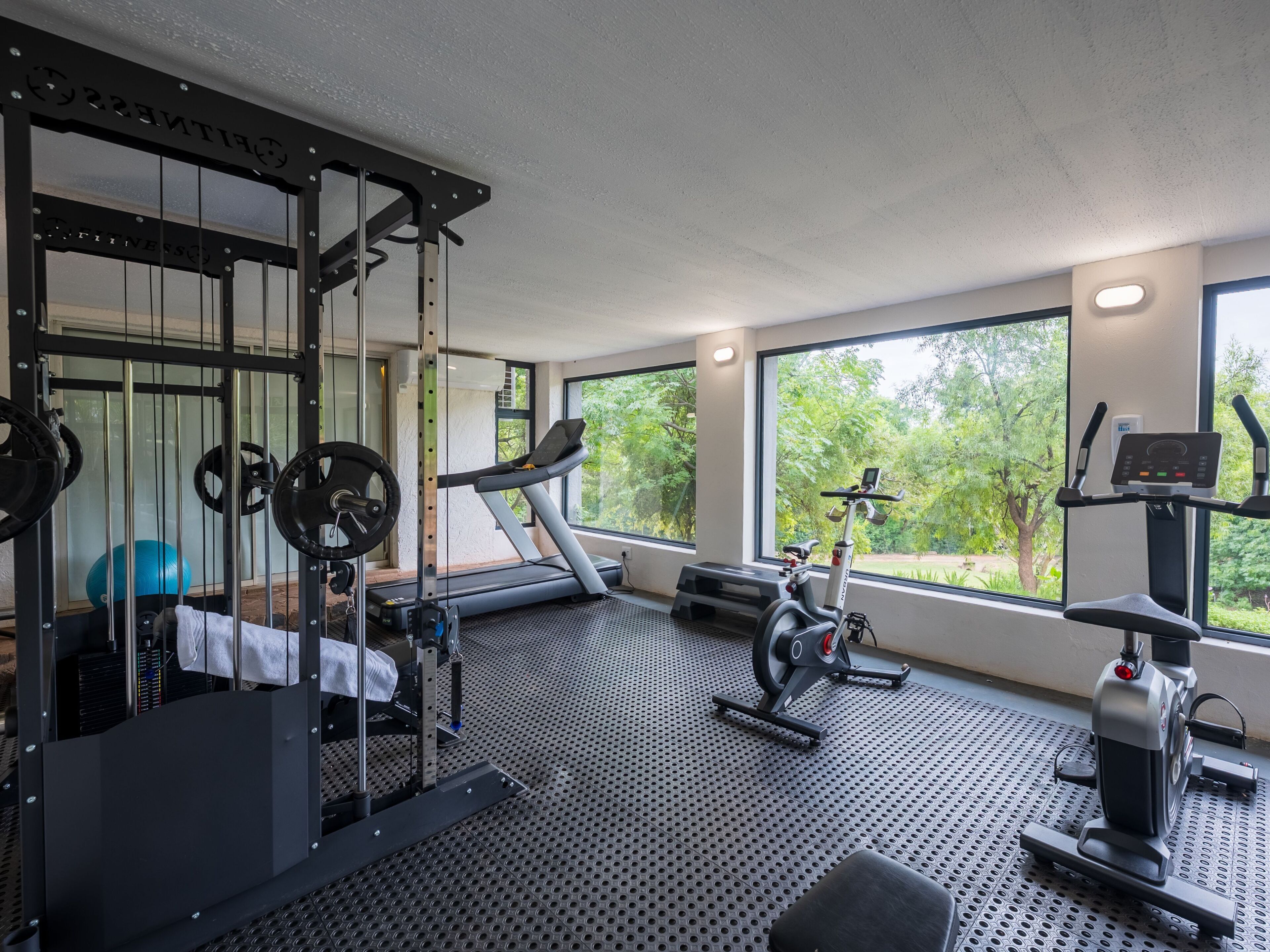 A gym with a treadmill, weight machine, and exercise bike, large windows with a view of trees, and a ceiling with recessed lighting.