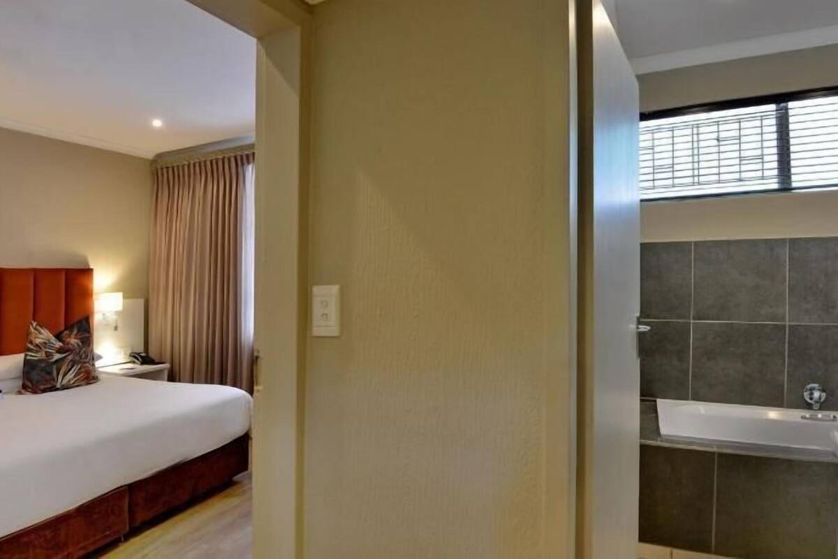 A hotel room with a bed, a nightstand, a lamp, a window with curtains, and a bathroom with a bathtub and tiled walls.