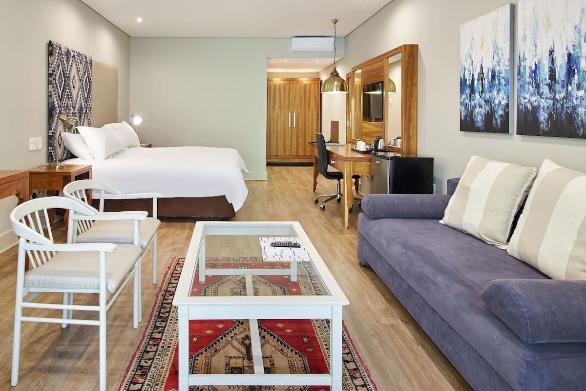 A hotel room with a bed, a sofa, a desk, and a chair. There is a rug on the floor and a painting on the wall.