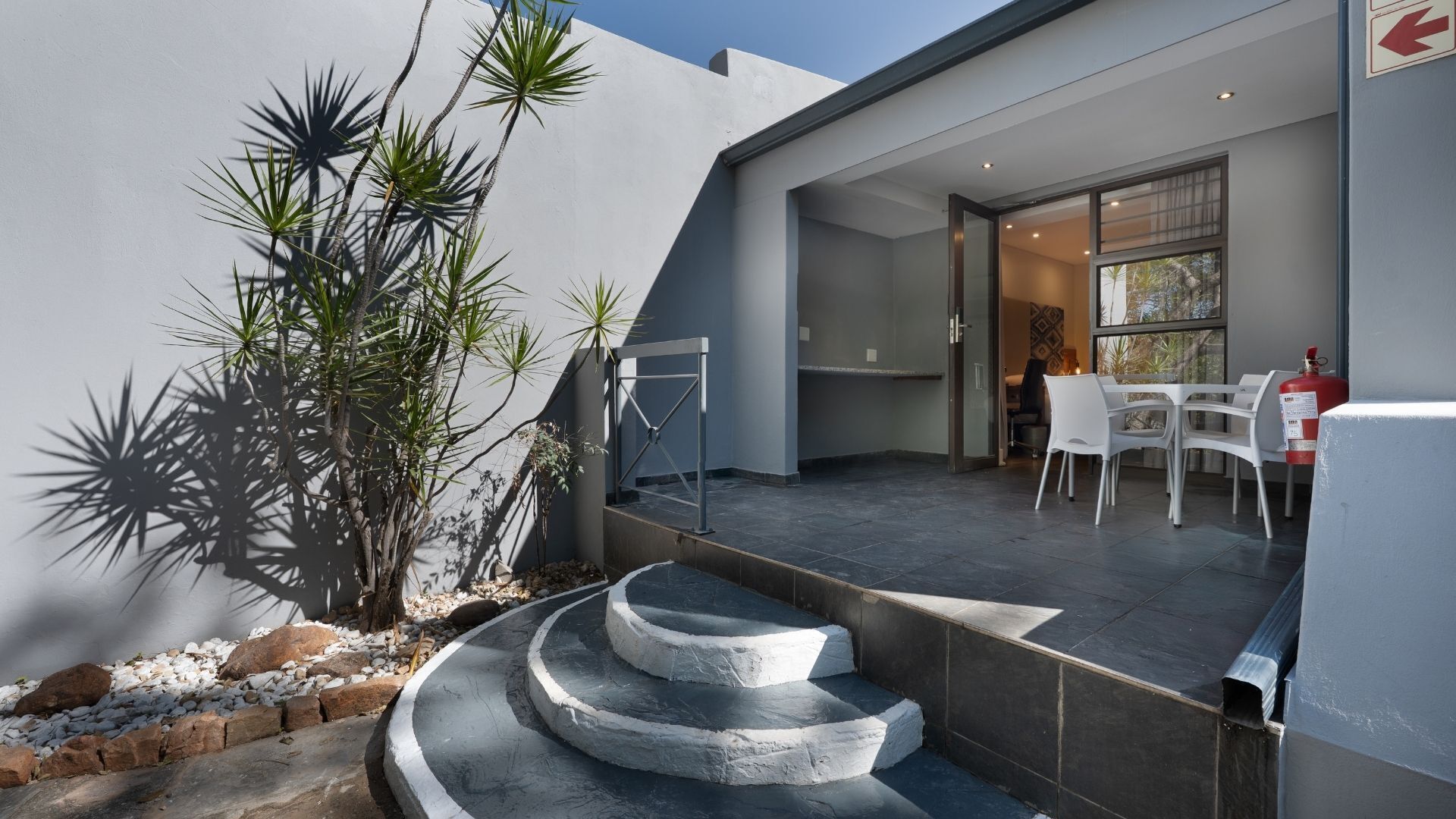 A modern outdoor patio with a staircase, white chairs, and a fire extinguisher.