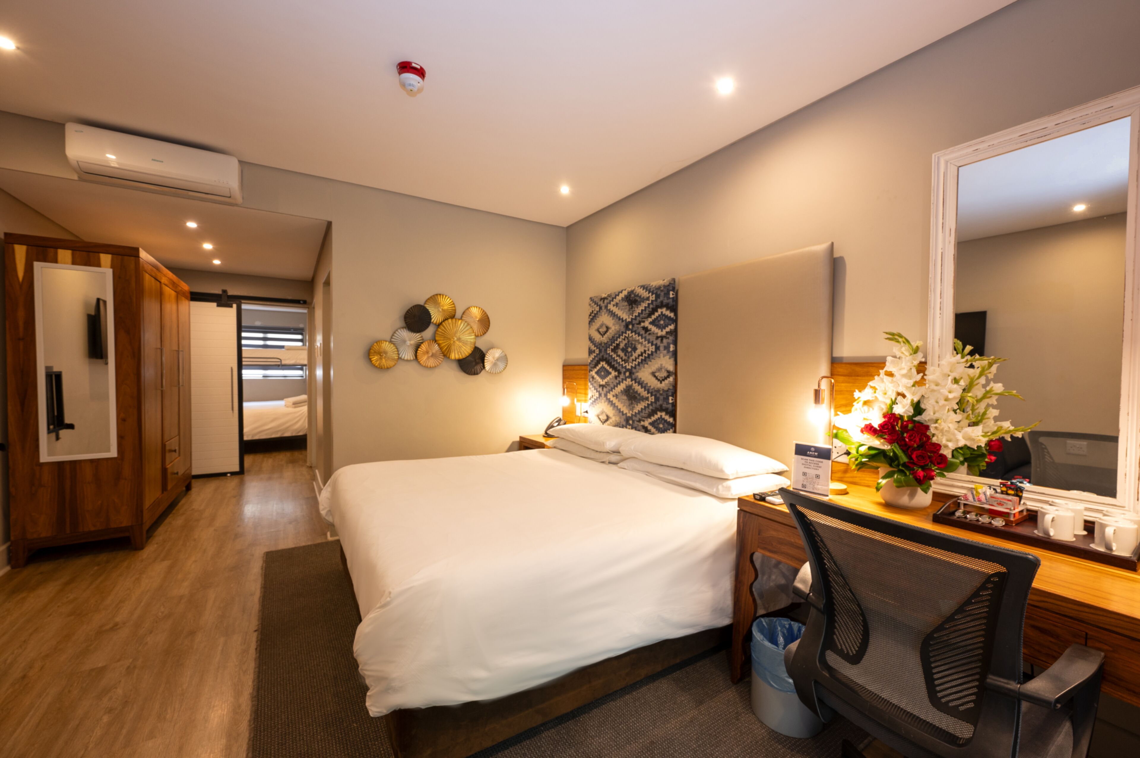 A hotel room with a bed, a desk with a chair, a mirror, and a wall-mounted air conditioner.