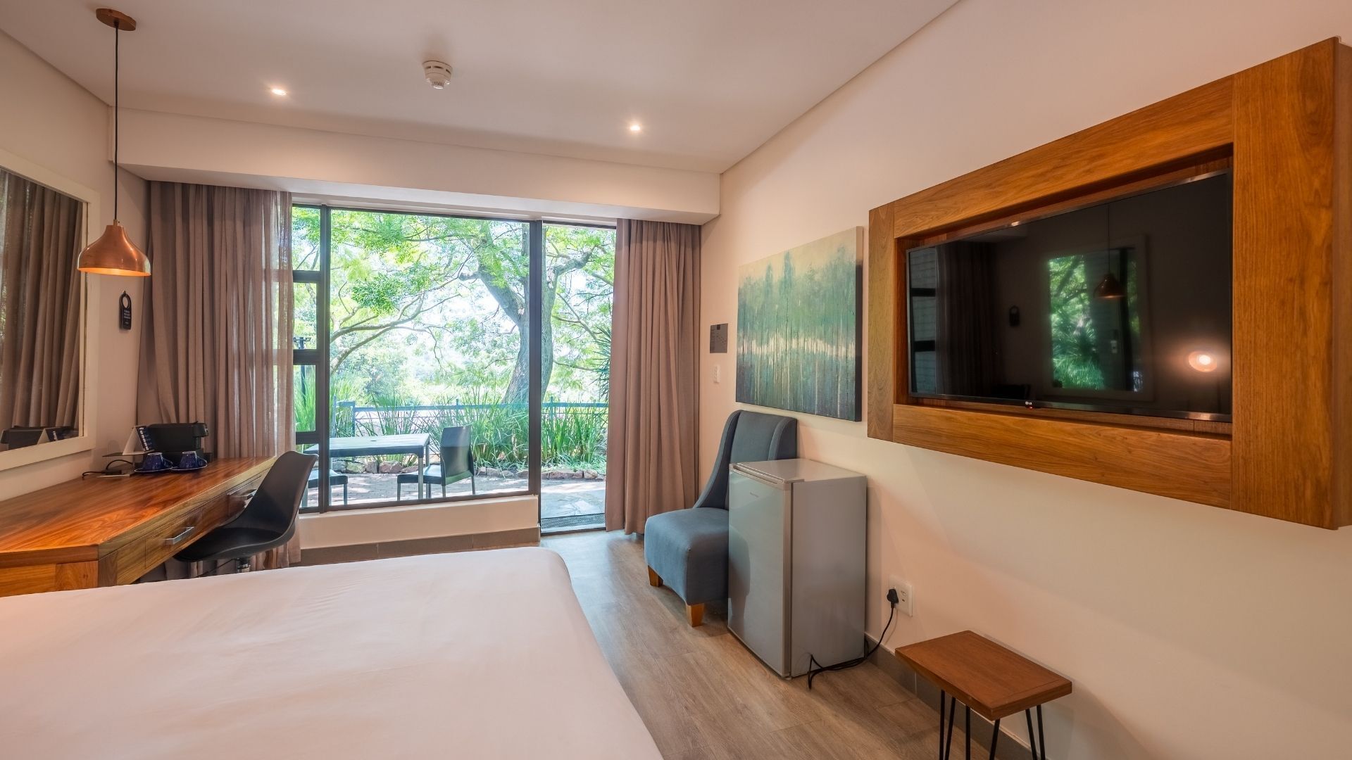 Habitació d'hotel amb llit, escriptori, cadira, televisió i un balcó que dona a arbres.