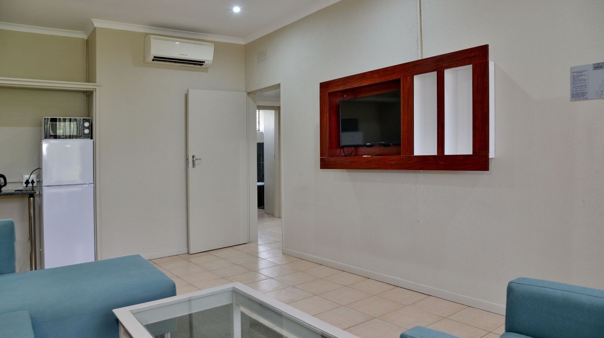 A living room with a glass coffee table, blue sofas, a wooden-framed window, and a white door leading to a kitchen with a refrigerator.