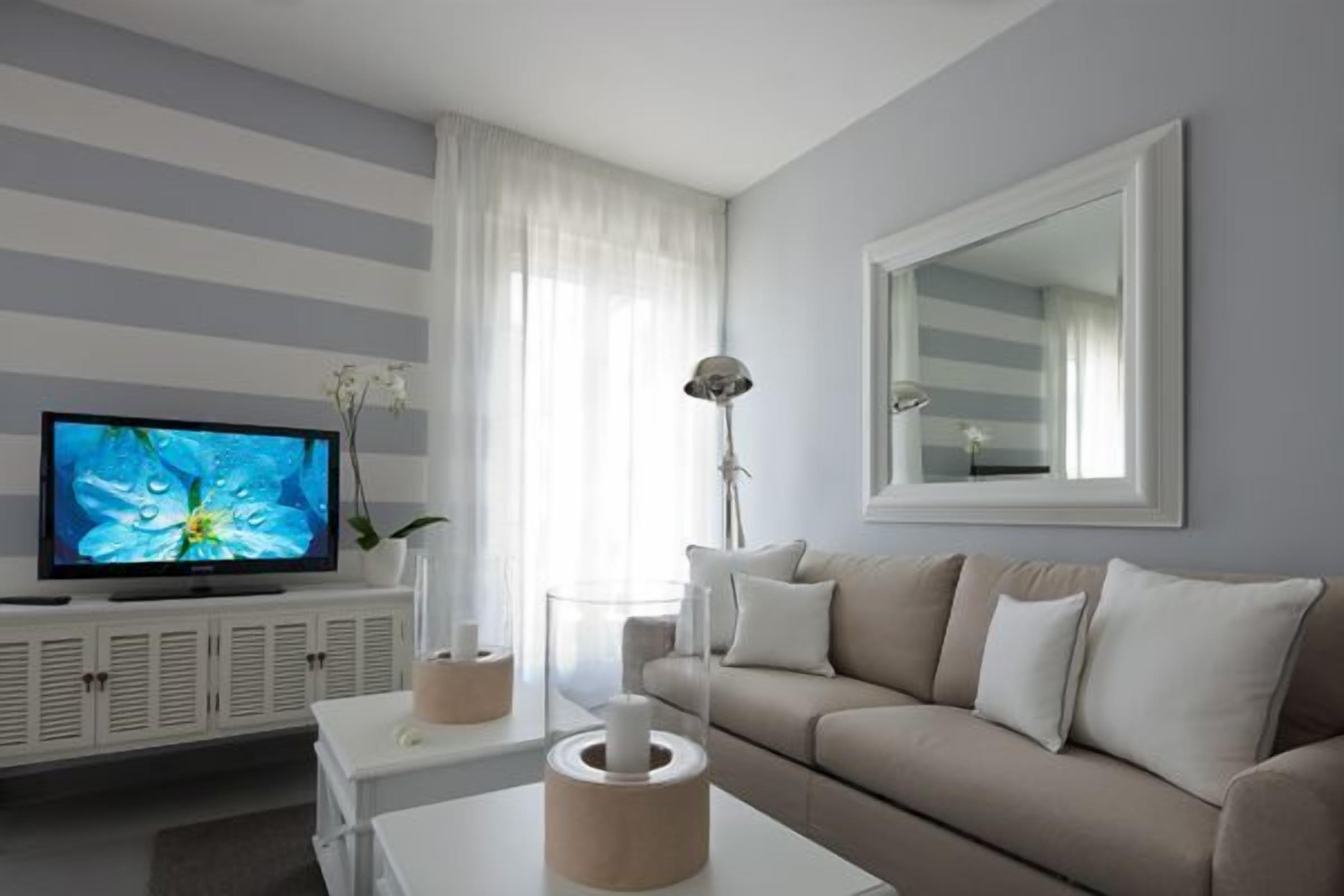 A modern living room with a flat-screen TV, a beige sofa, a white coffee table, and a large mirror on the wall.