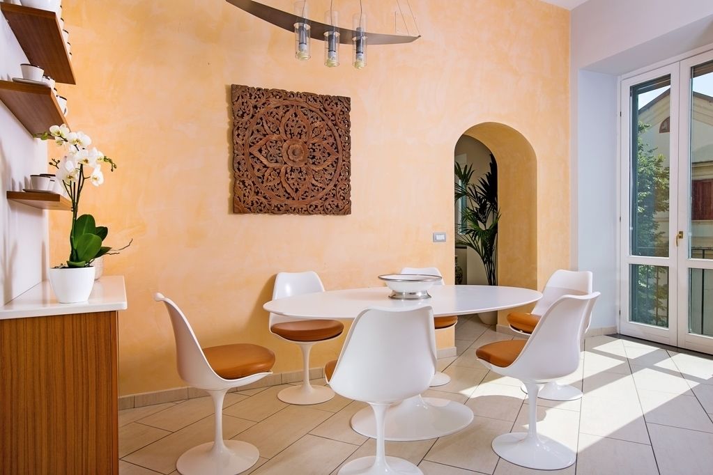 A modern dining area with a round table and white chairs, a decorative wall piece, and a potted plant.