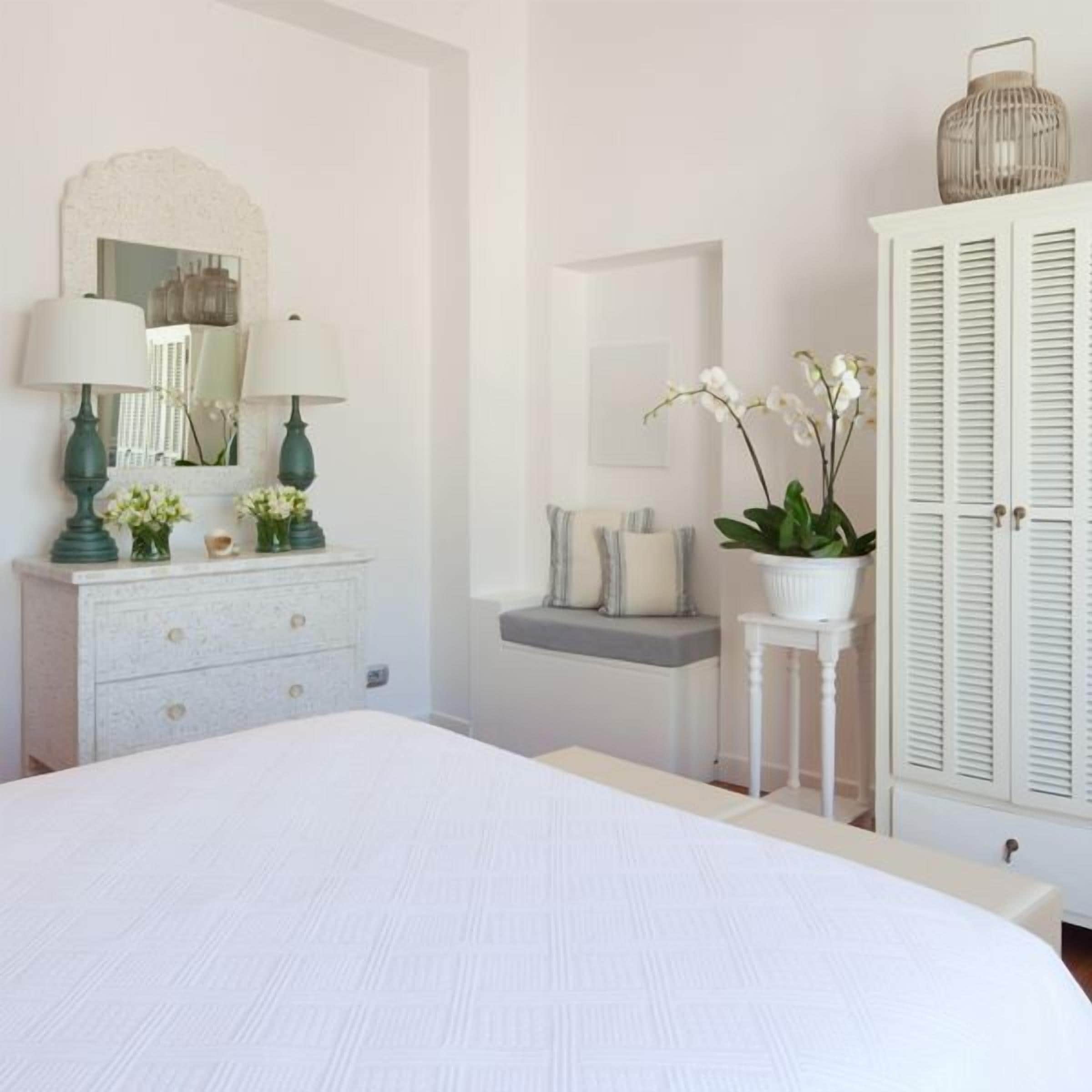 A bedroom with a large bed, a dresser with lamps, a mirror, a potted orchid, and a wardrobe.