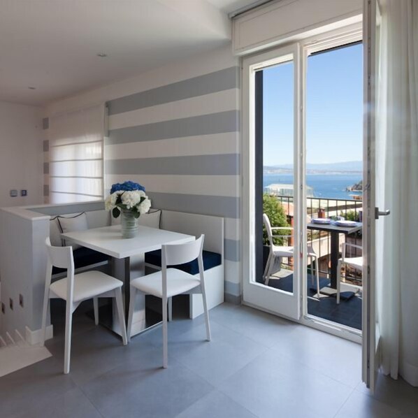 Une pièce moderne avec un mur rayé de blanc et de gris, une table blanche avec deux chaises et une vue sur la mer par une porte-fenêtre coulissante.