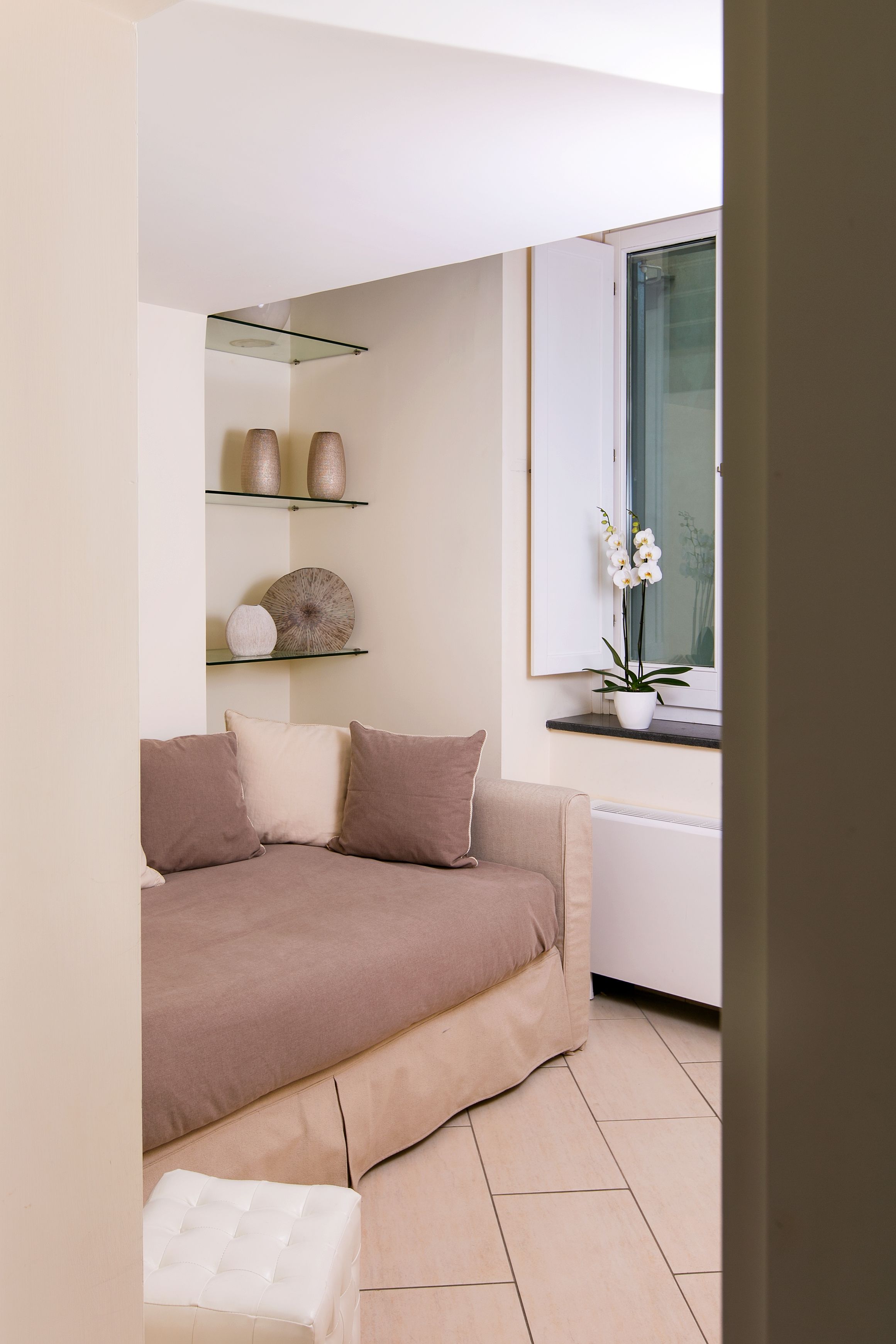 Un salon moderne avec un canapé beige, une étagère en verre ornée d’objets décoratifs et une fenêtre avec une orchidée en pot.