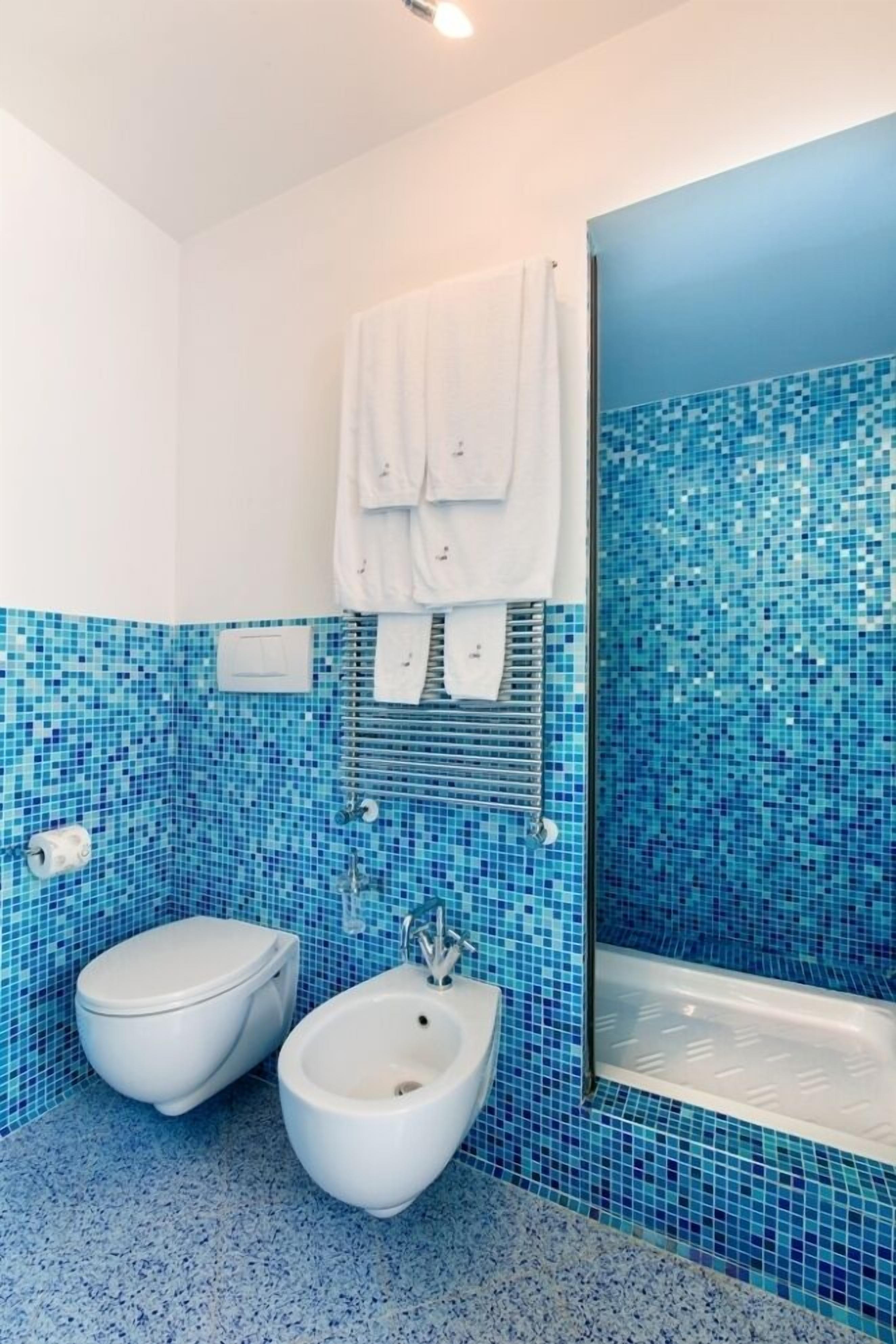 Une salle de bain avec des toilettes blanches, un bidet et une baignoire dont le mur est recouvert de carreaux de mosaïque bleus.