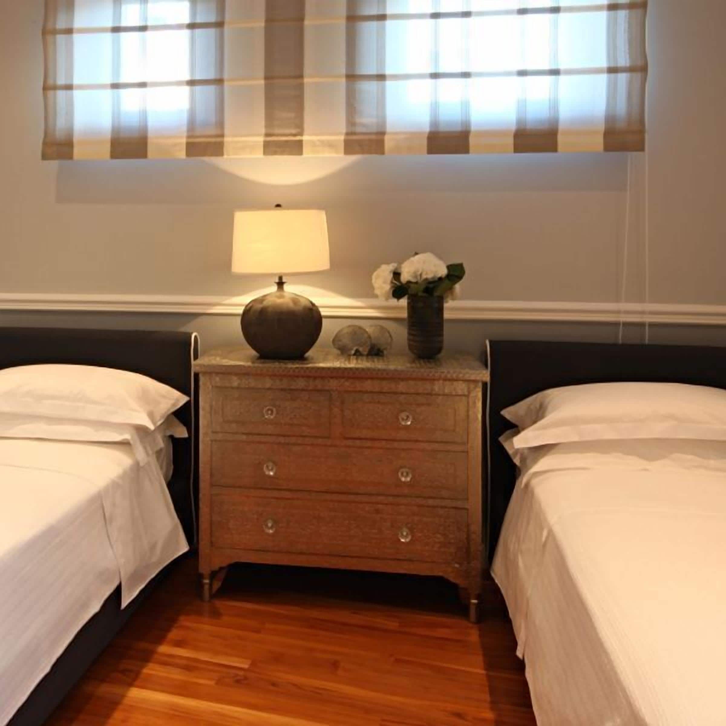 A bedroom with two beds, a nightstand with a lamp, and a vase with flowers.