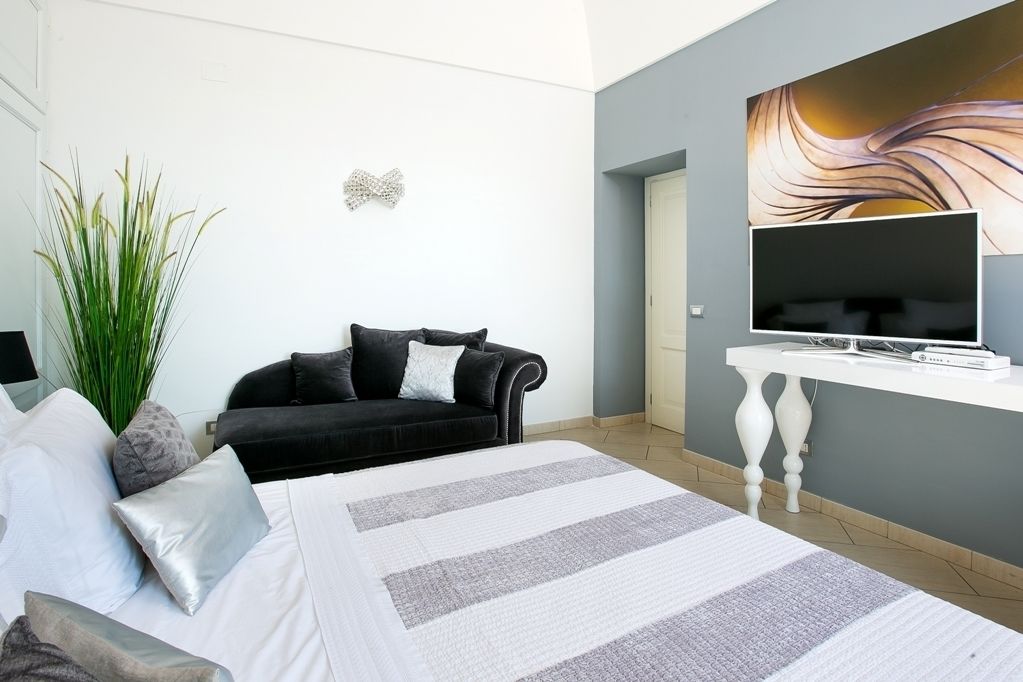 Une chambre à coucher moderne avec un lit, un canapé noir, une grande plante et un téléviseur posé sur une console blanche.