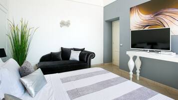 Une chambre à coucher moderne avec un lit, un canapé noir, une grande plante et un téléviseur posé sur une console blanche.