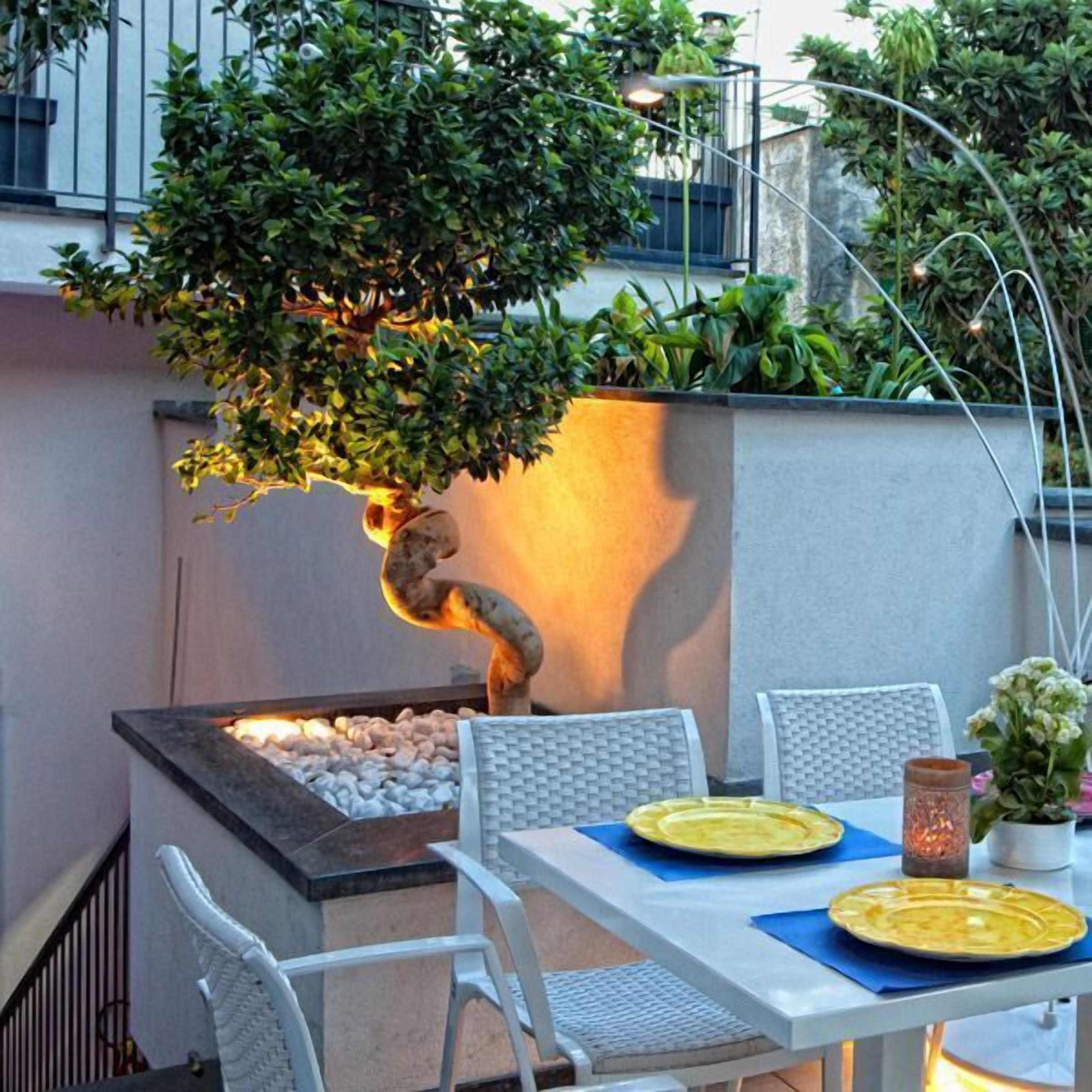 An outdoor dining area with a table set for two, surrounded by white chairs, a small fire feature, and a potted plant.