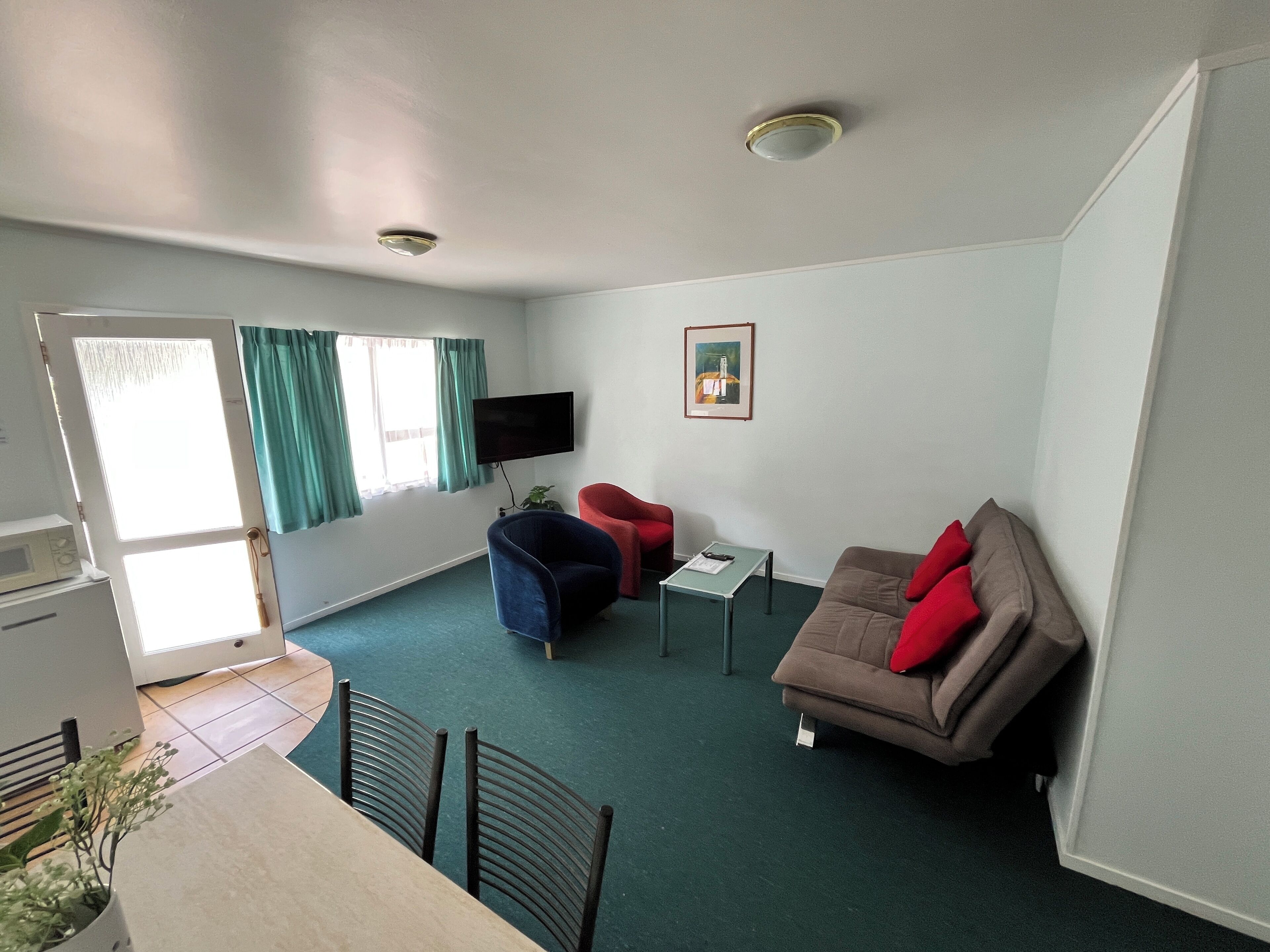 Ein gemütlicher Wohnraum mit einer Couch, einem Sessel, einem kleinen Tisch, einem an der Wand befestigten Fernseher und einem Fenster mit Vorhängen.