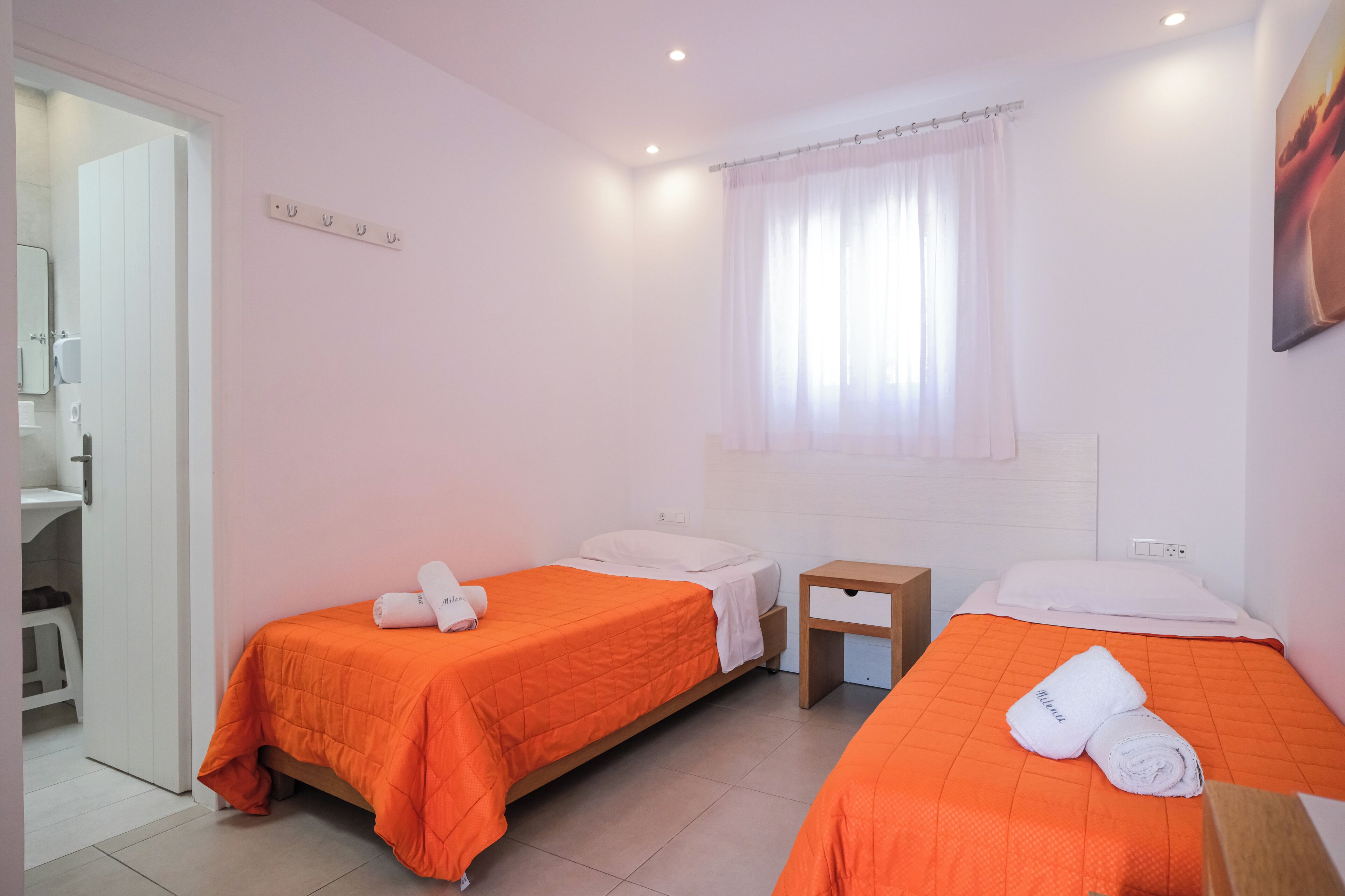 Habitación de hotel con dos camas, una mesita de noche de madera, una ventana con cortina blanca y una puerta blanca que da a un baño.