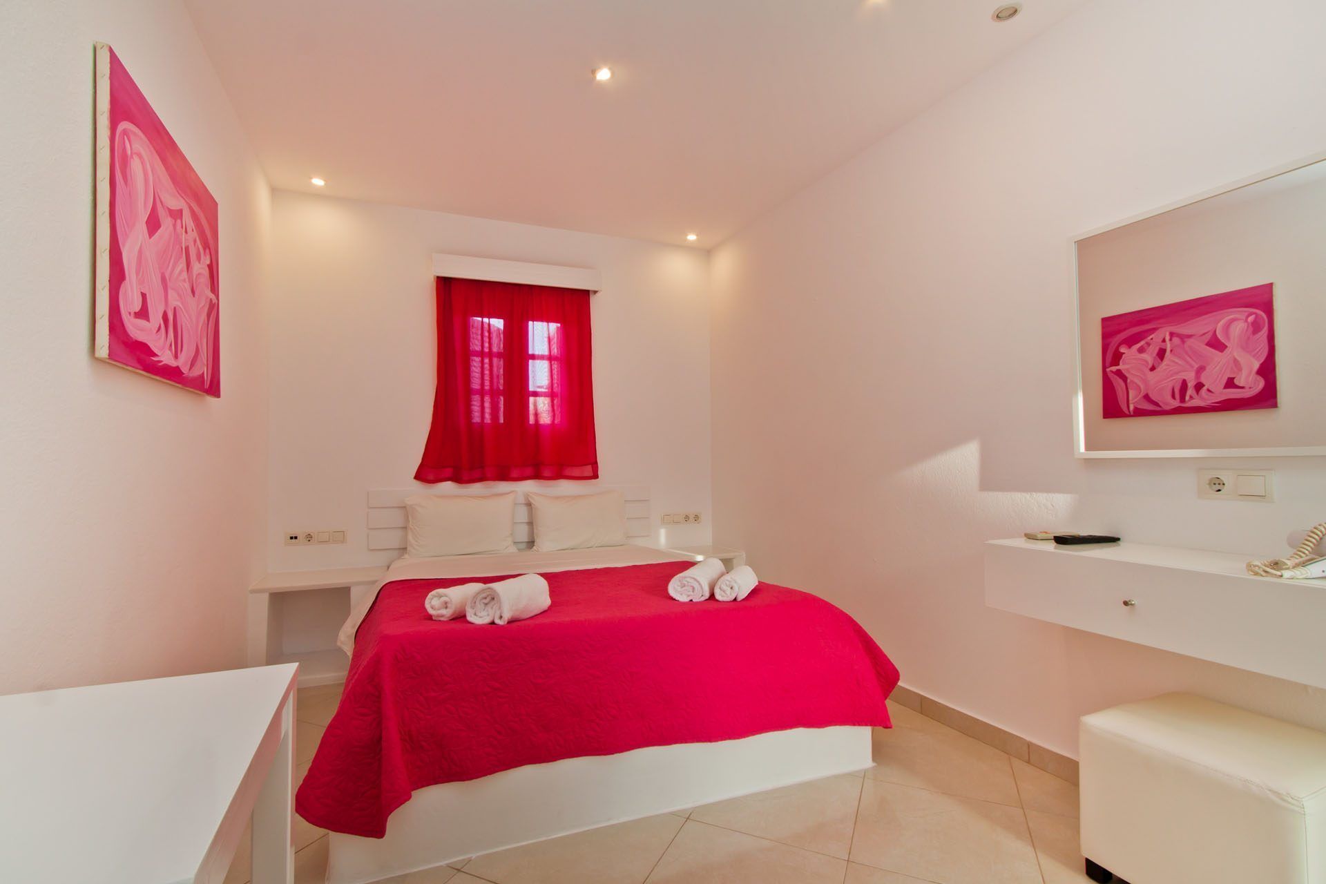 A hotel room with a bed, a desk, a chair, a window with red curtains, and artwork on the walls.